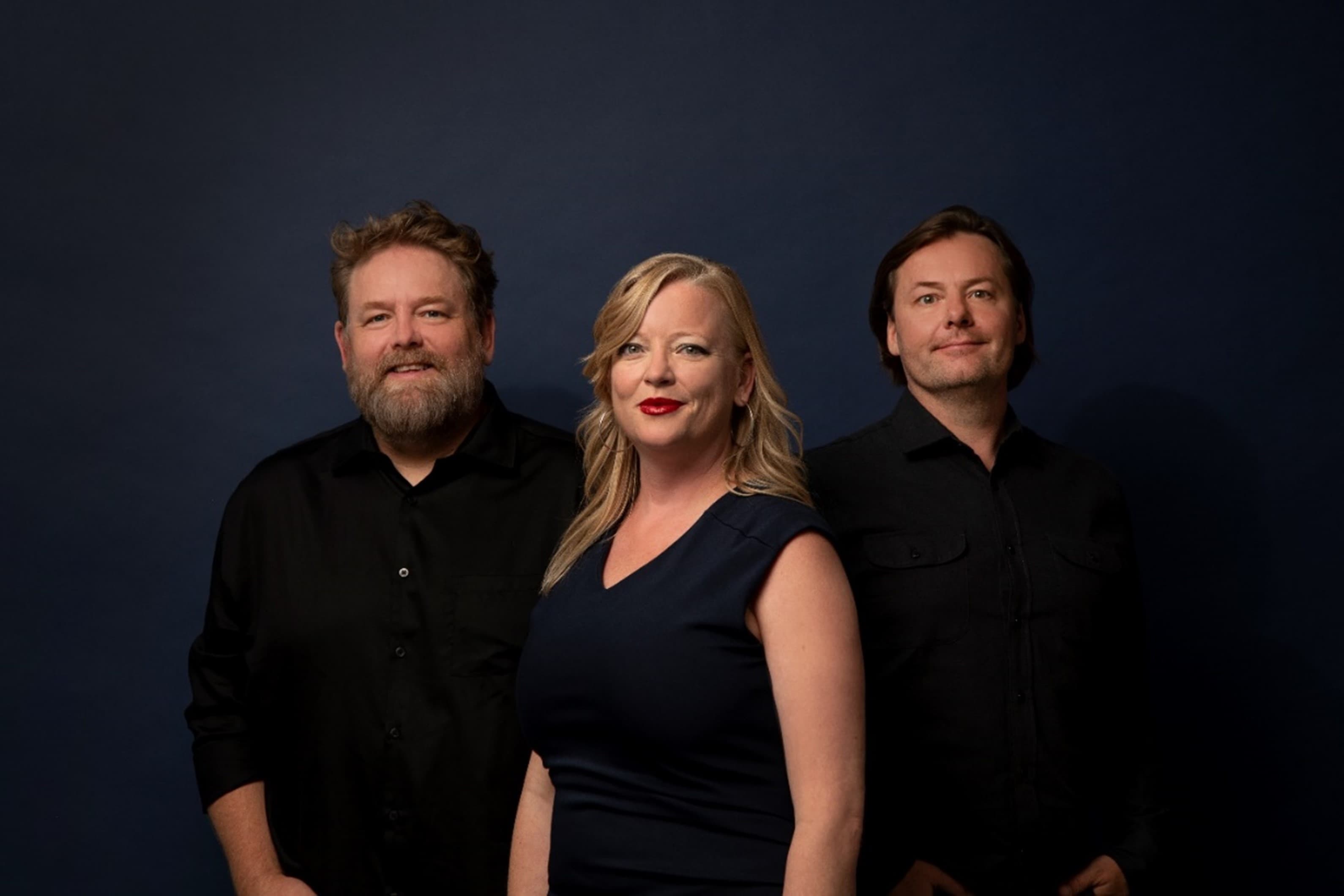 Image of the Rigadon trio, all wearing black and looking into the camera. They stand in front of a very dark greyish blue background.