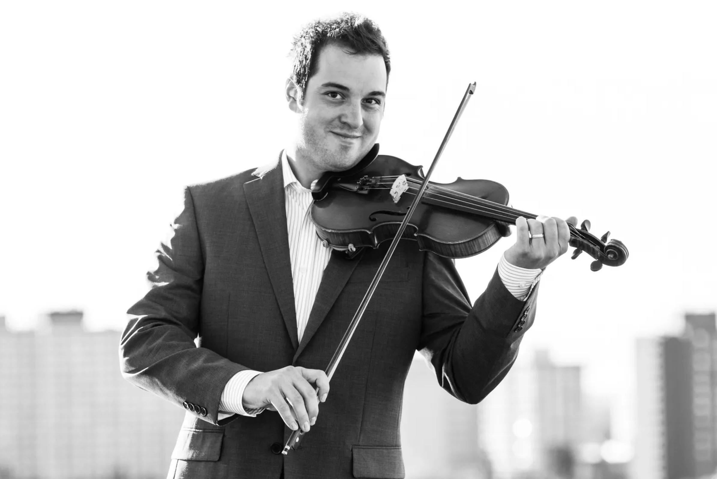 Black and white photo shows Daniel Gervais looking at the camera, standing in a suit-jacket, striped collared shirt, and playing a violin.