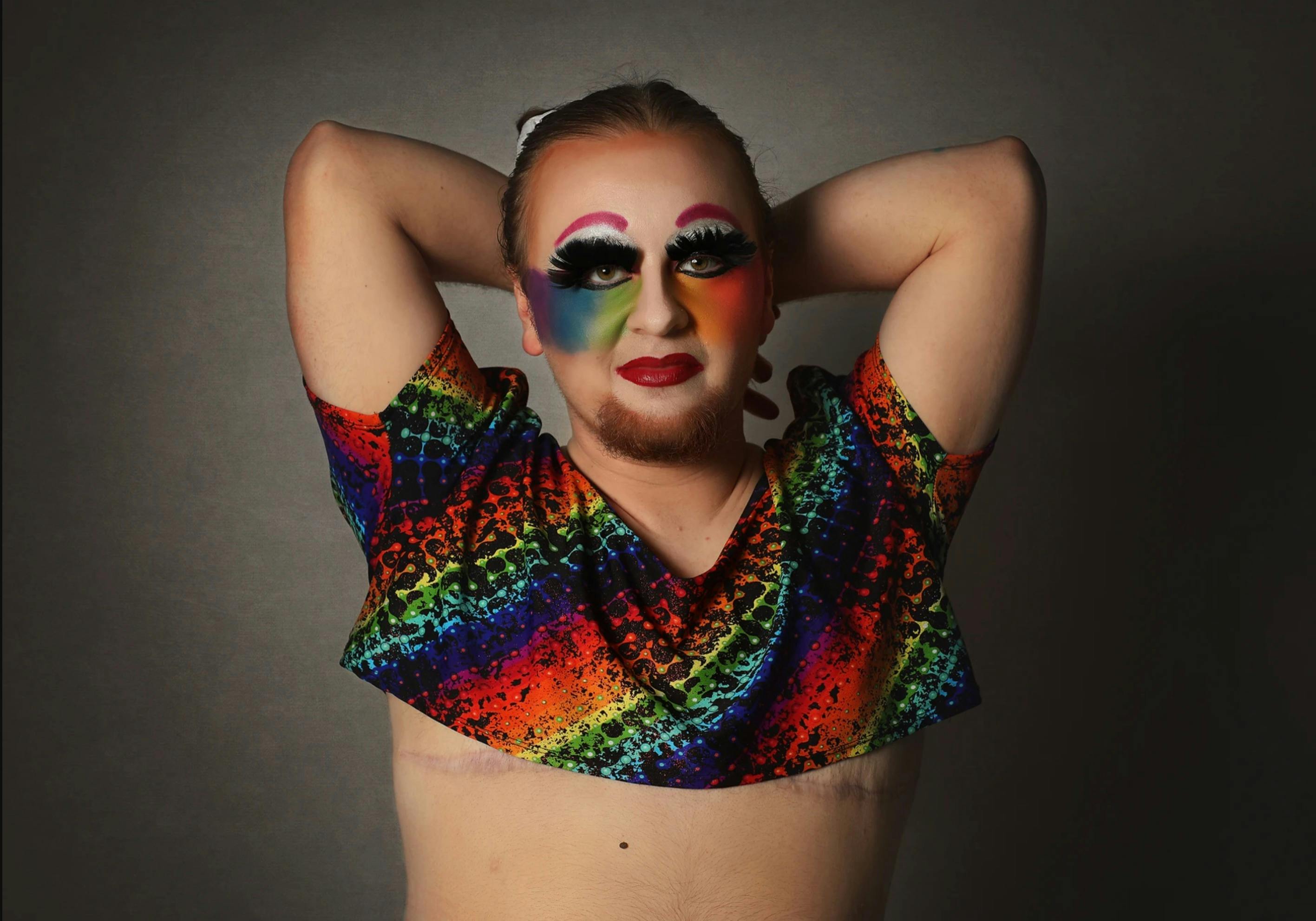 Coco LaQueer looks at the camera with full lashes and pink eyebrows, wearing rainbow face makeup and a colourful rainbow crop top.